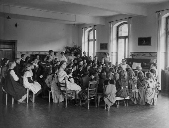 Black and white picture. Many children and some women are sitting on chairs in a room.