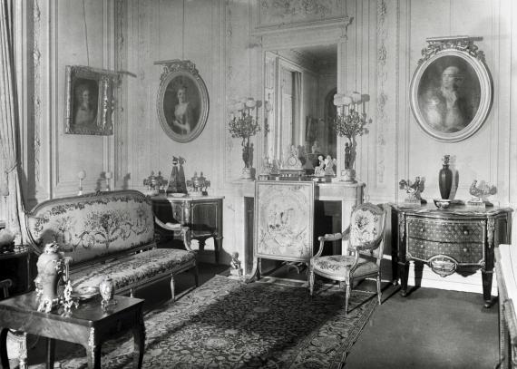 Black and white photograph of Hermine Feist's rococo-style drawing room. In addition to seating furniture by the fireplace, there are large, ornate chests of drawers and a variety of objets d'art. Portraits from the 18th century adorn the walls.