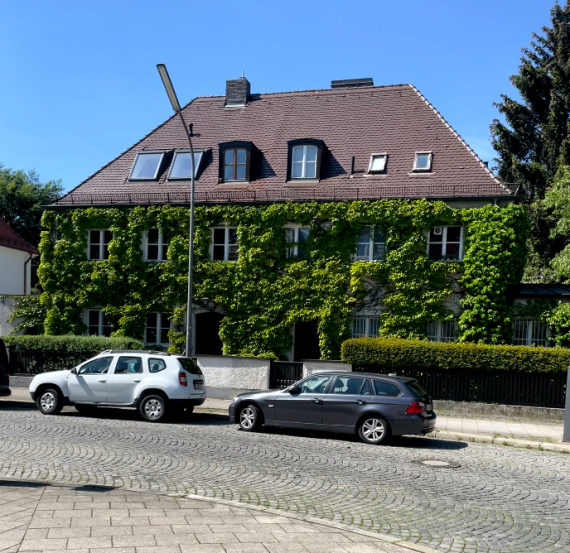 Frontal view of a detached house, overgrown with ivy. The street can be seen in front of it.