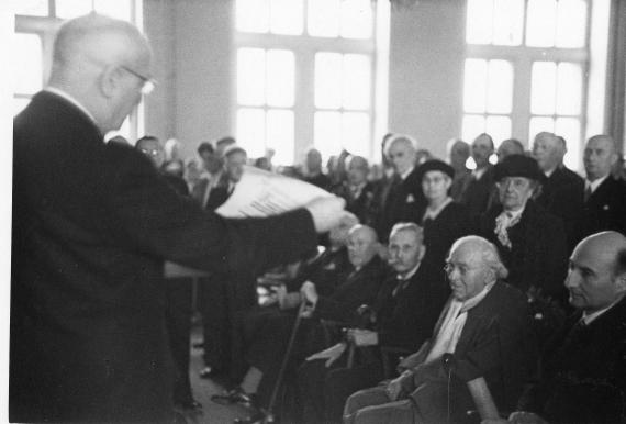 Black and white picture. Hall with many people. A speaker in the foreground.