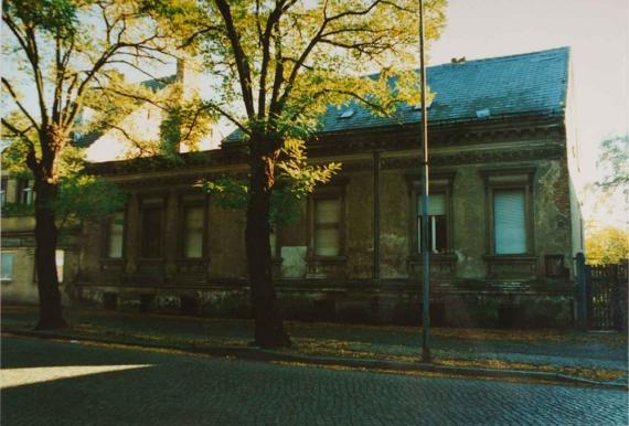 The picture shows an older two-storey house that is in a neglected condition. The façade is dark and heavily weathered, with peeling paint and visible damage. The windows have lowered shutters and are surrounded by decorative frames. There are several large trees in front of the house, some of whose leaves cast shadows on the building. The sidewalk in front of the house is paved, and a street lamp stands to the right of the picture. The roof of the house is dark and also in poor condition.