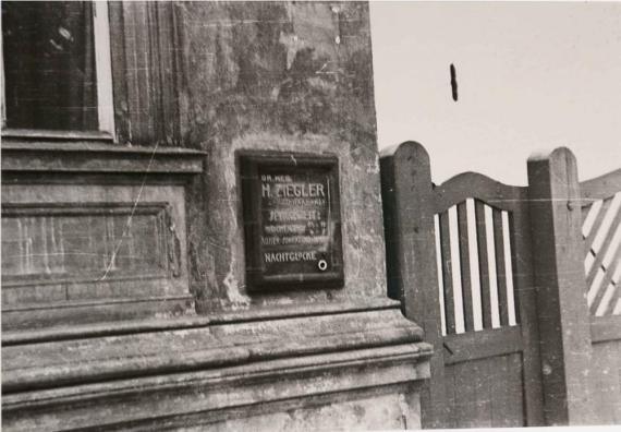 Das Schwarz-Weiß-Foto zeigt eine Nahaufnahme eines Schildes an einer Hauswand neben einem Fenster. Das Schild trägt die Aufschrift "Dr. med. H. Ziegler" und weist darauf hin, dass es sich um eine Praxis handelt, vermutlich für einen Arzt. Unter dem Namen steht "Facharzt für Innere Medizin und Lungenheilkunde". Rechts vom Schild ist ein hölzernes Tor zu sehen. Die Fassade des Gebäudes wirkt abgenutzt und zeigt deutliche Altersspuren.