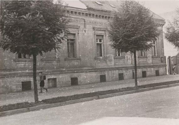 Das Schwarz-Weiß-Foto zeigt ein älteres Gebäude mit einer stark abgenutzten Fassade. Es ist zweistöckig mit dekorativen Stuckarbeiten und großen, rechteckigen Fenstern. Die Fenster haben heruntergelassene Rollläden. Vor dem Gebäude verläuft ein gepflasterter Gehweg, auf dem ein Kind läuft. Es gibt mehrere Bäume entlang des Gehwegs, die teilweise das Gebäude verdecken. Das Bild wirkt historisch und zeigt Spuren von Abnutzung und Alterung am Gebäude.