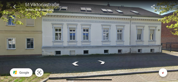 Das Haus in der Viktoriastraße 52 in Velten, Brandenburg, ist ein zweistöckiges Gebäude mit einer weißen Fassade, dekorativen Stuckelementen und Fenstern mit Umrahmungen. Das Dach ist mit roten Ziegeln gedeckt und hat mehrere Dachfenster. Das Gebäude wirkt gepflegt und hat einen klassischen, historischen Stil.