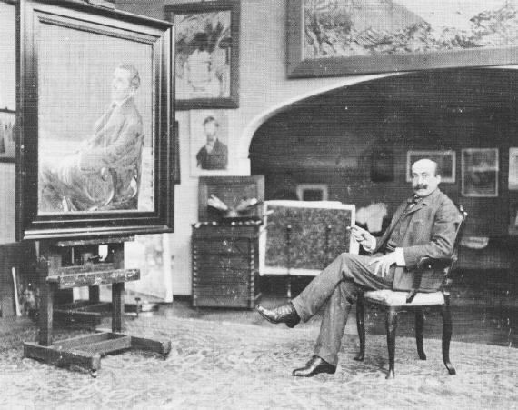 A black and white photograph shows the painter Max Liebermann sitting on an armchair in a suit. He is in his spacious, salon-like studio, which is hung with framed works on the walls and covered with a large carpet. On a large easel stands a huge, framed painting that is around 1.50 m high and wide.