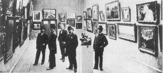 A black and white photograph shows an exhibition space. Paintings hang on the wall in two rows, one above the other in decorative frames. In the center of the photo are 5 men in suits, one of whom is Max Liebermann