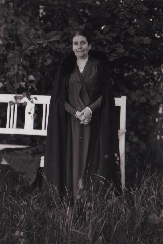 A black and white photograph shows an elderly woman in dark clothing in a garden