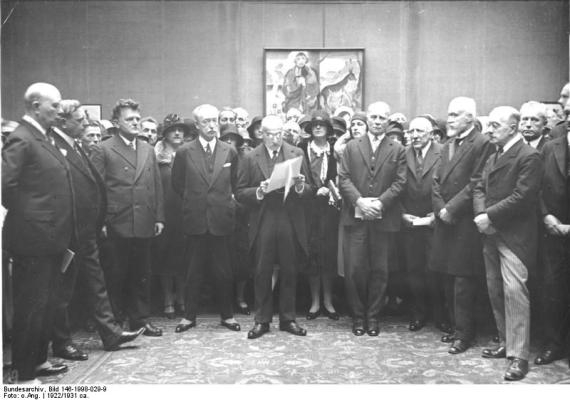 A black and white photo shows Liebermann with a speech script in an exhibition room. Many listeners stand in a semicircle around him