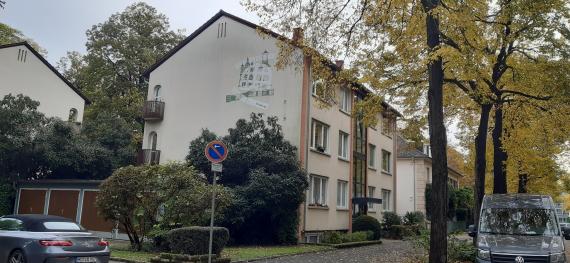 Seitenansicht auf ein Gebäude, daneben der Gehweg und Bäume, und parkende Autos