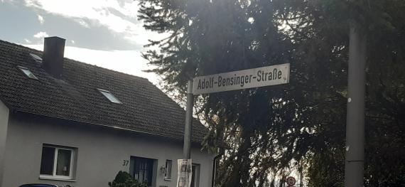 Straßenschild, im Hintergrund links ein Haus, rechts ein Baum