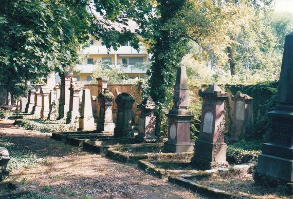 A row of old gravestones. Surrounded by trees. A new house in the background.