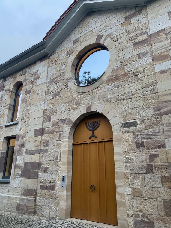 Das Bild zeigt das Portal der Felsberger Synagoge im Jahr 2023. Im Fenster über der großen Holztür spiegelt sich die Felsburg. 