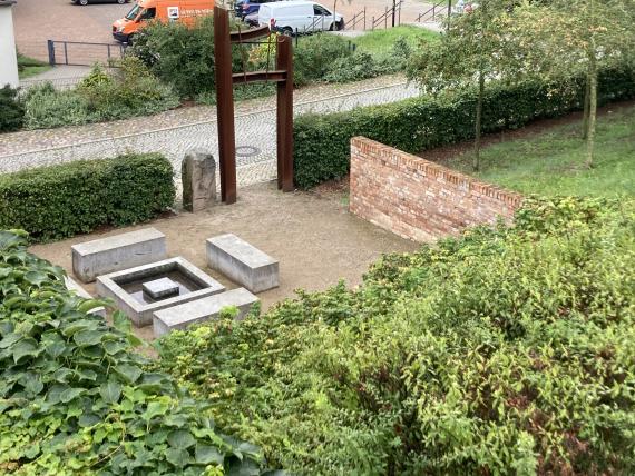 View of the memorial from the Jewish steps. At the top center of the picture is a broken, rust-red steel frame with a golden Star of David. In front of it, stones with pools of water symbolizing a miquveh. On the right, part of the 12 fruit trees.