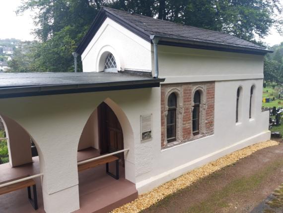 The tahara hall with gabled roof and the slightly lower open porch are plastered white. The original exposed brickwork is visible in two of the four windows. There is an information board in the middle of the building complex.