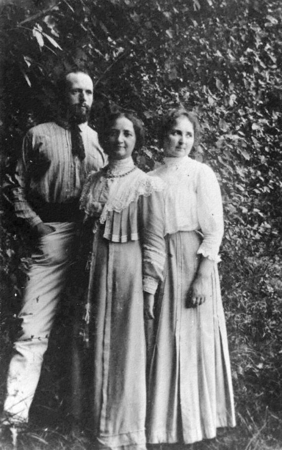 Two women and a man pose in typical 19th century bourgeois clothing in front of a leafy hedge