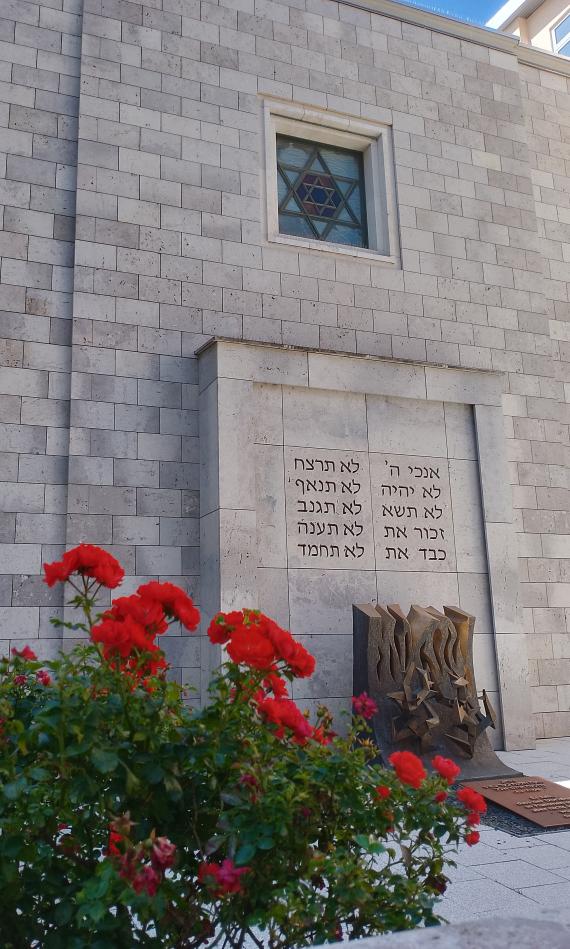 Im Hintergrund zu sehen die schlicht gehaltene Fassade der Stuttgarter Synagoge aus grau-cremfarbenem Travertin-Stein. In der Mitte ist die Rückseite des Tora-Schreins zu sehen. Darauf die Zehn Gebote bzw. deren Anfangsworte in hebräischen Buchstaben. Oberhalb ist ein quadratisches Fenster mit David-Stern zu sehen. Vor dem Gebäude ist eine aus Bronze gefertigte Skulptur von Roda Reilinger, die einen brennenden Dornbusch zeigt.