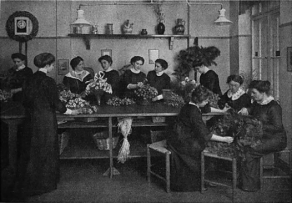 Aus der Schule für Blumenschmuck von Franziska Bruck in Berlin ist der Untertitel einer Aufnahme von Becker & Maass zum Zeitungsartikel Eine Schule für Blumenschmuck (Autorin: Ola Alsen) The black and white photograph shows 10 young women sitting and standing around a large table and tying various bouquets of flowers. In the background you can see a shelf with various flower vases. They are students of Franziska Bruck's school for flower arranging. They are dressed in the fashion of the time. They are all wearing a long dress with a white collar. Their hair is pinned up.