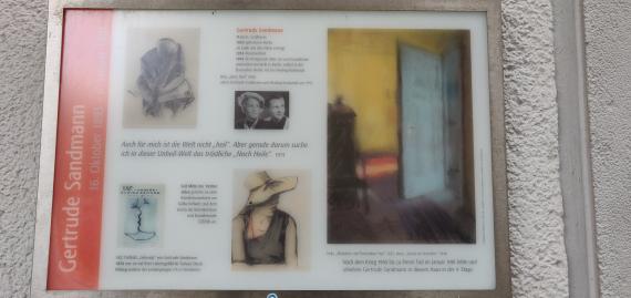 Das Foto zeigt eine Gedenktafel mit Fotografien von Gertrude Sandmann und ihrer Freundin Hedwig Koslowski. Drei Grafiken sind abgebildet. Sie zeigen zwei Frauengestalten und das Sonnenlicht in einem Flur.
