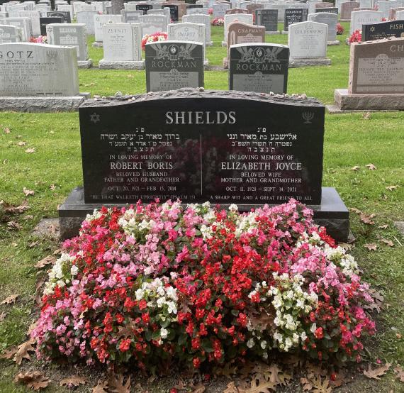 Ein Grabstein auf einem Friedhof. Auf dem Grab ist ein Blumenbeet. 