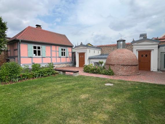 Links im Bild steht das Synagogendienerhaus aus grauem Fachwerk und rosafarbenem Putz. Rechts daneben schließt sich das jüdische Ritualbad an. Es besteht aus unterschiedlichen Gebäudeteilen. Den Hintergrund bildet ein rechteckiger Querriegel mit einem nach Westen abfallenden Pultdach, der an den Enden von zwei Risaliten eingefasst ist. Die nach Westen weisenden Eingangsfronten wurden tempelartig gestaltet. Asymmetrisch rechts davor versetzt befindet sich ein backsteinerner Kuppelbau mit Laterne, welcher das Tauchbad überdacht.