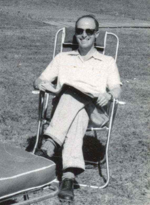 Schwarz-Weiß Bild. Ein Mann mit Sonnenbrille sitzt entspannt in einem Gartenstuhl und lächelt.