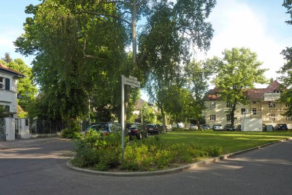 Starßenschild des Johanna-und-Willy-Brauer-Platz in Berlin Karlshorst. Es ist ein grüner wiesiger Platz mit Wohnhäusern und ein paar Bäumen im Hintergrund.