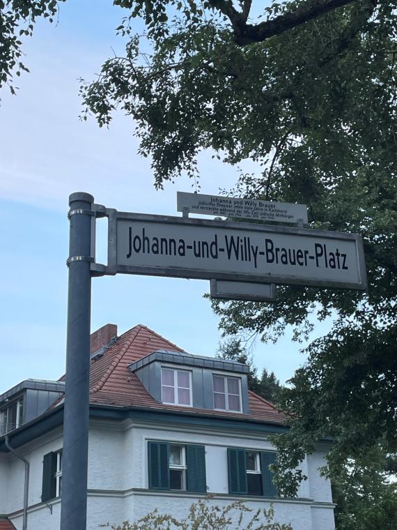 Nah fotografiertes Straßenschild des Johanna-und-Willy-Brauer-Platz mit näheren Informationen zu dem jüdischen Ehepaar. Im Hintergrund sieht man einen blauen Himmel und ein weißes Wohnhaus mit grünen Fensterläden und rotem Dach sowie einen Baum.