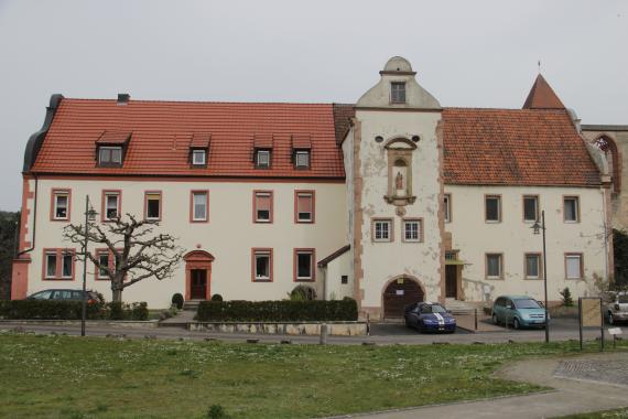 Das Foto zeigt ein ehemaliges Kloster mit beiger Wand und rotem Dach. Man sieht den Haupteingang  und ein paar Autos stehen auf dem Parkplatz davor. 