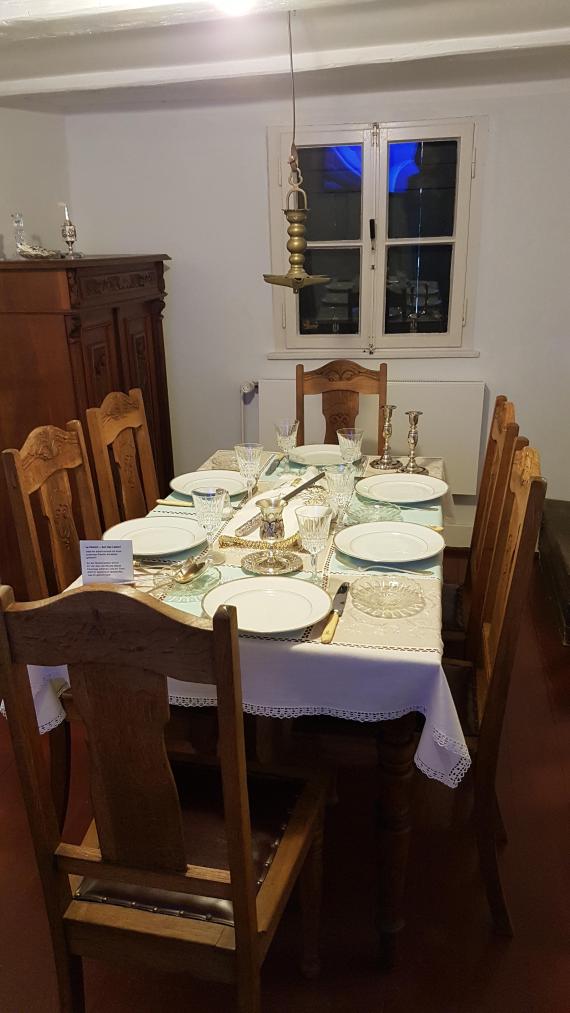 In the parlor, the table is festively set to celebrate Shabbat. There are 6 chairs around a table. An old oil lamp hangs above the table. Candlesticks to welcome Shabbat are also placed on the table. The spice jar stands on a vertiko, an old sideboard. Through the window facing the street, you can see that it is getting dark.