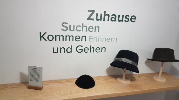 A kippah, an old women's hat and a men's hat lie on a shelf in the hallway. On the wall are the words, home, coming and going, remembering and searching.