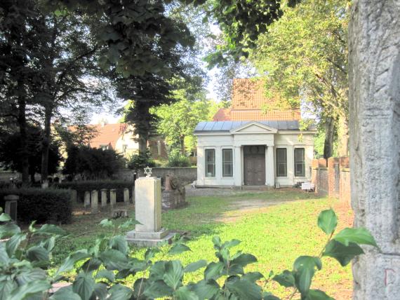 Der Blick fällt auf die Zeremonienhalle im klassizistischen Stil. Davor befindet sich ein Gedenkstein für die jüdischen Opfer des Nationalsozialismus.