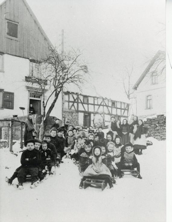 In the foreground of the almost 100-year-old photo, you can see all the children of the village lined up like a school class with sledges. Adult women (mothers?) are to the left and mostly to the right of the group. In the background you can see the gable of the house and a woman dressed in black in the doorway. It could be Mrs. Gottschalk.