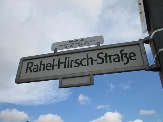 Das Bild zeigt ein Straßenschild der Rahel-Hirsch-Straße. Auf dem Schild steht ein kleineres Schild, worauf steht: Rahel Hirsch Erste Medizinprofessorin in Preußen geb. 1870 gest. 1953