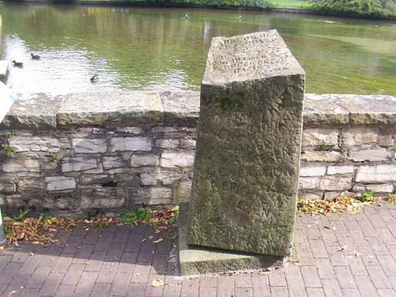 Das Foto zeigt den Gedenkstein für Jenny Aloni, der neben einem kleinen Gewässer und vor einer kleinen Mauer in einem Park steht. Der Gedenkstein ist aus Naturstein Material und ein nicht rechteckiger Quader, der vermutlich 1 Meter hoch ist und 30cm breit und lang. Auf dem Stein sind verschiedene Wörter graviert, die sich auf dem Foto nicht gut lesen lassen und er ist schon ein wenig mit Moos bewachsen. 