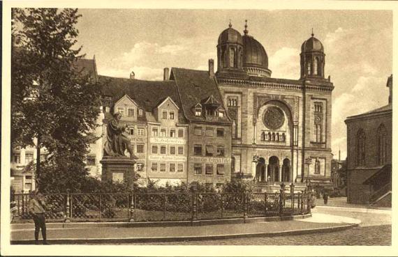 The square in front of the synagogue with trees and sculpture on the left. Synagogue with two towers and three domes