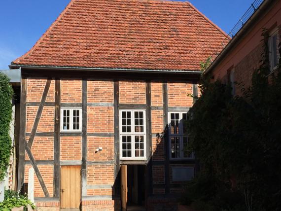 Half-timbered building with fields of red brick and white lattice windows. The one-story building has a simple red tile roof. In front of the building on the left there are two gravestones.