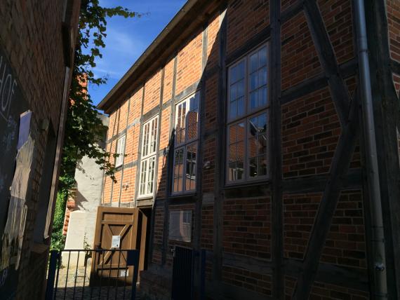 Half-timbered building with fields of red brick and three large windows with white coffered frames. The view from the street is into a small courtyard from which one enters the synagogue. The entrance door is open.