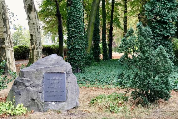 On a meadow and near a sidewalk is a memorial stone about 1.5 meters high and 1.5 meters wide. In the background there are large trees. On the stone is a large memorial plaque with the text: "Here was the synagogue of the Jewish community Hohenschönhausen 1935-1938".