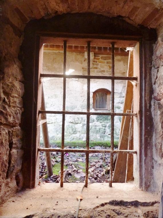 Das Bild zeigt ein vergittertes Fenster in einer Bruchsteinmauer von Innen nach Außen. Man sieht auf die direkt benachbarte Außenwand der Mühle.