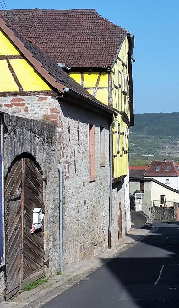 Mazzenbäckerei und Geschäft für Landesprodukte Jakob Hirschenberger. Gelbes Fachwerkhaus, die Bäckerei befand sich im kleinen Nebengebäude, die Wohnung von Jakob Hirschenberger und das Geschäft für Landesprodukte befand sich im großen Haupthaus. Im Hintergrund Blick auf das Maintal.