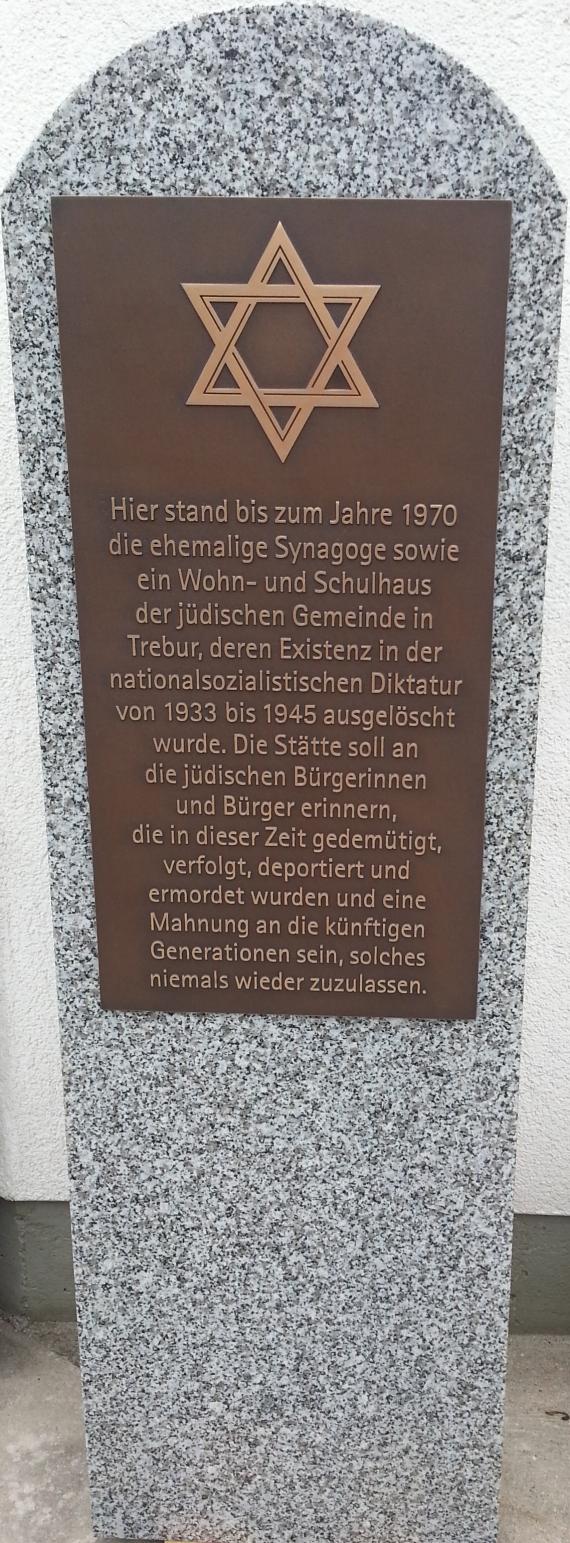 Große, länglicher Granitstein. Auf der oberen Hälfte ist mittig eine Eisenplatte angebracht, auf der ein Davidstern sowie ein Erinnerungstext sowie eine Mahnung zu lesen sind. 