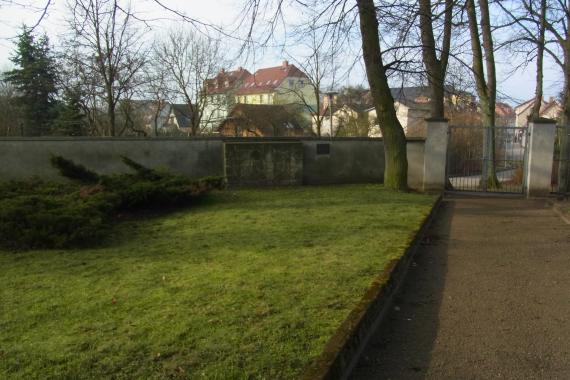 On the left lawn with bushes, behind it plastered wall with walled fragments of the destroyed Jewish gravestones and a small bronze memorial plaque. Next to the lawn path to the cemetery entrance and tall trees.