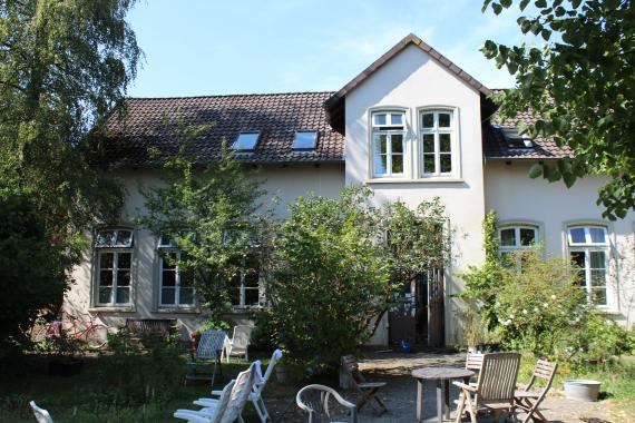 Front view of a white one-story house with a slanted roof. In the foreground a lawn with garden tables and garden chairs.