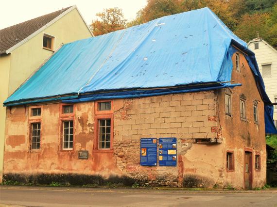 Die ehemalige Synagoge Laudenbach, es sind deutliche Schäden und Reparaturversuche am Mauerwerk zu sehen. Das Dach ist mit einer blauen Plastikplane großflächig abgedeckt.