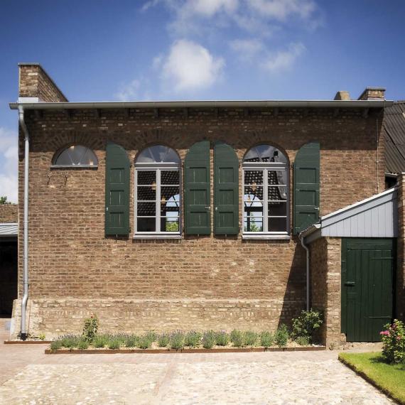 Small building with two large, high windows. The windows have large green shutters on both sides. The windows are still finished with a semicircular window at the top. To the left is a third semicircular window. Below this semicircular window there is only brickwork. In front of the building there is a small bed with lavender. In front of the building, on the right side, there is a small privy with a green door.